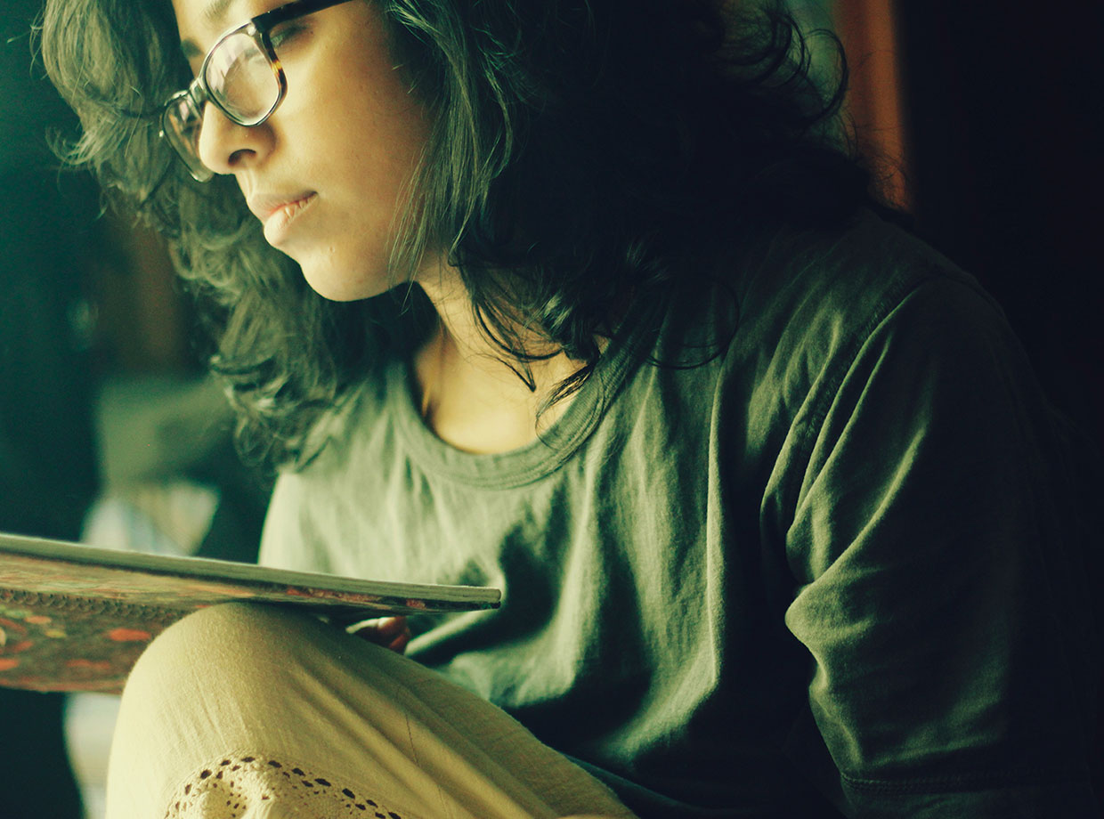 A person with long, wavy hair and glasses is sitting and reading a book from Oxford University Press. They are wearing a casual green shirt and light-colored pants. The scene has a soft and introspective ambiance, with warm lighting illuminating their face from the side.