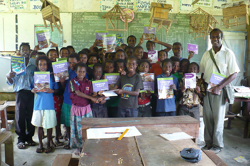 A group of smiling children and adults in a classroom, proudly holding up language workbooks. The classroom has wooden benches, desks, and educational materials hanging from the ceiling. The chalkboard in the background displays various educational notes.